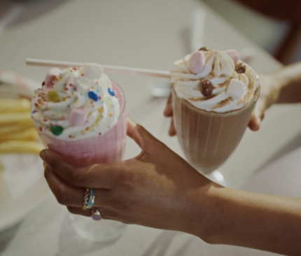 Hands holding two milkshakes with a burger and chips in the background